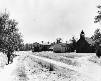 Three wooden structures in a row at the intersection of two dirt roads. A lone bicyclist rests in the foreground. 